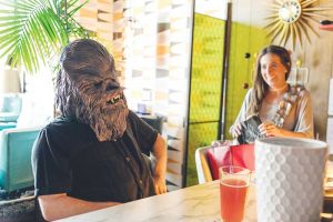 Someone sitting at a coffee counter while wearing a Chewbacca mask as a woman looks on